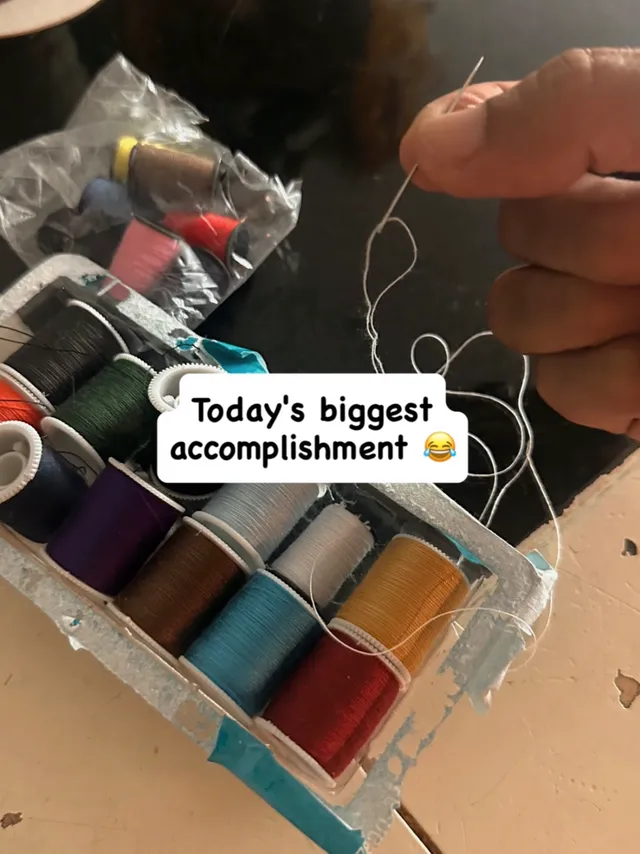 A hand threading a needle above a box of coloured cotton spools